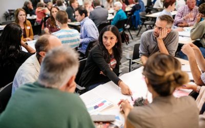 At a crowded community forum, a woman points to a chart paper, surrounded by other people at a table.