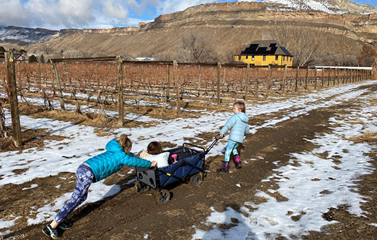 Two young girls pushing a smaller girl in a cart across a dirt field covered with snow, and a mountain in the background